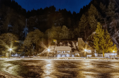 A cozy lodge illuminated at night, surrounded by trees and a waterfall in the background, with a wet, reflective parking area.