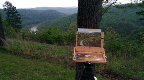 A painter's easel with a landscape painting overlooks a lush green valley and winding river in the distance.