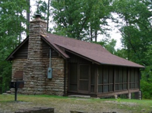 A rustic stone cabin surrounded by trees, featuring a chimney and a screened porch.