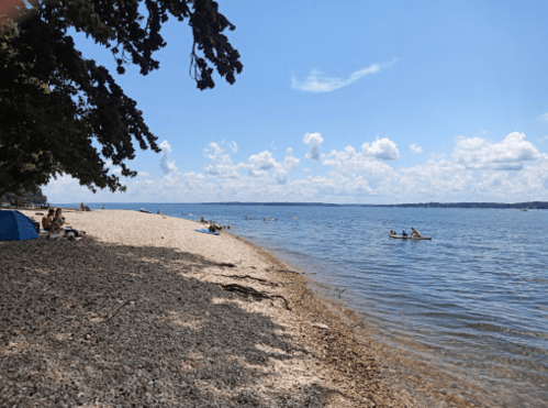 A sunny beach scene with people relaxing on the shore and kayaking in the calm water under a blue sky.