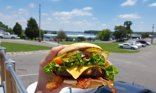 A hand holding a loaded cheeseburger with bacon, lettuce, tomato, and pickles, with a scenic lake view in the background.