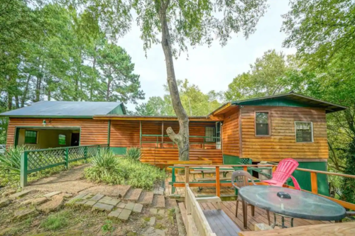 A rustic wooden cabin surrounded by trees, featuring a deck with outdoor seating and a stone pathway.