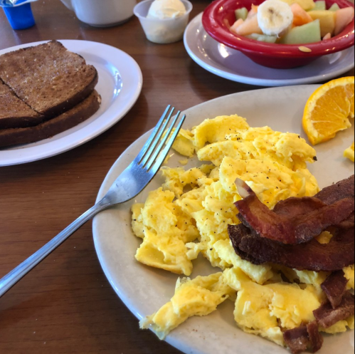 A plate of scrambled eggs, bacon, and a slice of orange, with toast and a fruit bowl in the background.