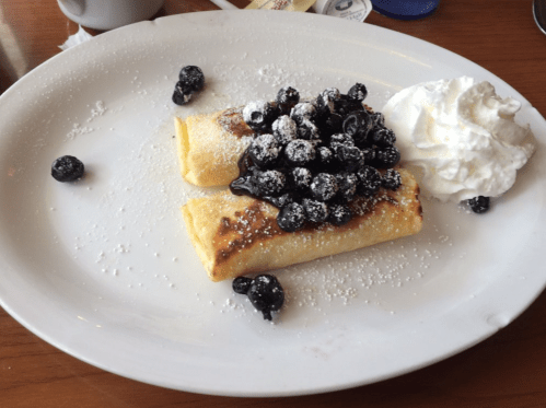Two crepes topped with blueberries and powdered sugar, served with a dollop of whipped cream on a white plate.