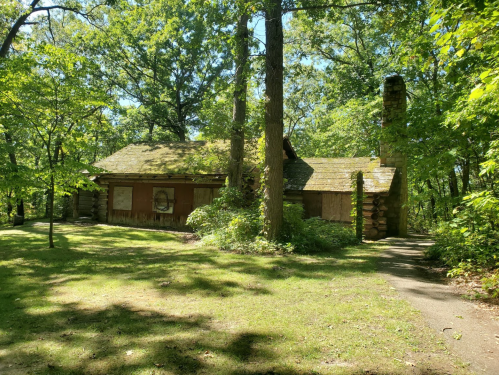 Abandoned log cabin surrounded by trees, with boarded windows and a stone chimney, set in a lush green landscape.