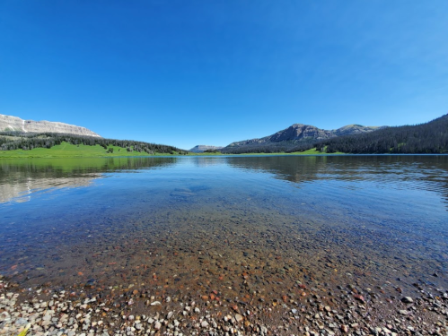 A serene lake surrounded by green hills and mountains under a clear blue sky, with a rocky shoreline in the foreground.