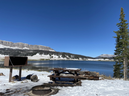 A snowy lakeside scene with a picnic table, fire pit, and mountains under a clear blue sky.