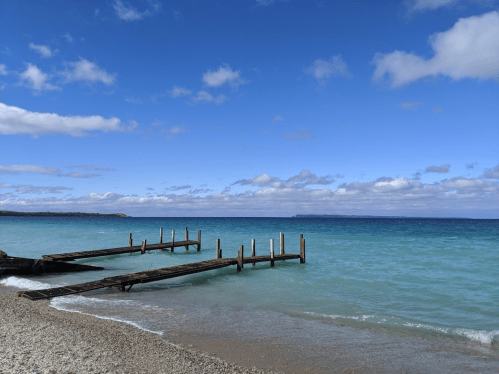 A serene beach scene featuring a wooden dock extending into calm turquoise waters under a partly cloudy sky.