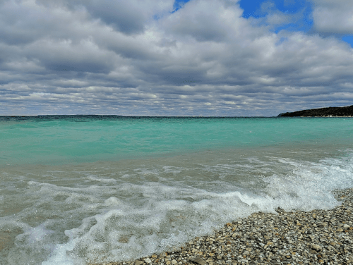 A tranquil beach scene with gentle waves lapping at a pebbly shore under a cloudy sky.