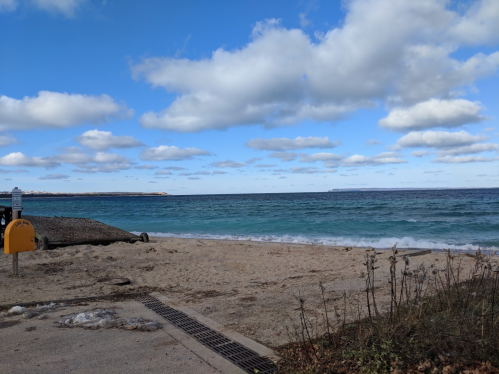 A sandy beach with gentle waves, under a blue sky filled with fluffy clouds. A distant island is visible on the horizon.