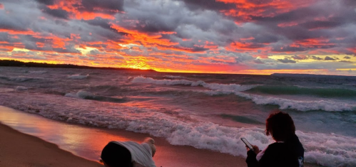 Two people on a beach at sunset, with vibrant orange and purple clouds reflecting on the water and waves.