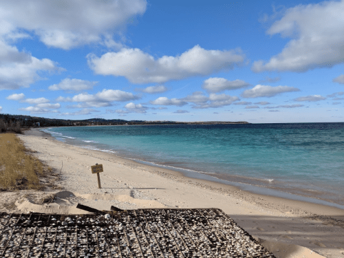 A serene beach scene with gentle waves, sandy shore, and a clear blue sky dotted with clouds.