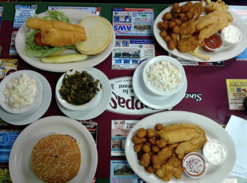 A spread of Southern-style dishes including fried fish, hushpuppies, coleslaw, greens, and a burger on a colorful table.