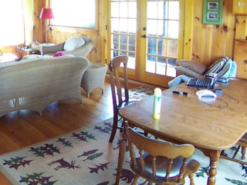 Cozy wooden interior with a table, chairs, and a couch, featuring large windows and a patterned rug.