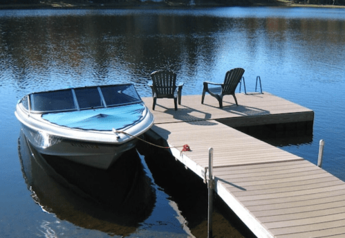 A boat is docked at a wooden pier with two chairs beside it, reflecting on calm water.