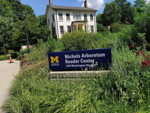 Sign for Nichols Arboretum Reader Center in front of a white house, surrounded by greenery and trees.