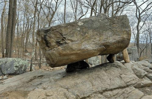 A large, balanced rock sits atop a smaller stone, surrounded by bare trees and rocky terrain.