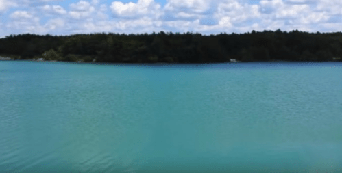 A serene lake with turquoise water, surrounded by green trees and a blue sky dotted with fluffy white clouds.