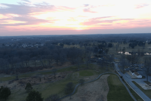 Aerial view of a landscape at sunset, featuring trees, a golf course, and a small pond in the distance.