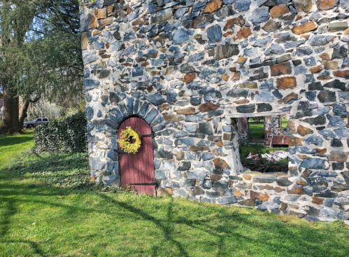 A stone building with a red door and a wreath, surrounded by green grass and trees. A window is visible nearby.