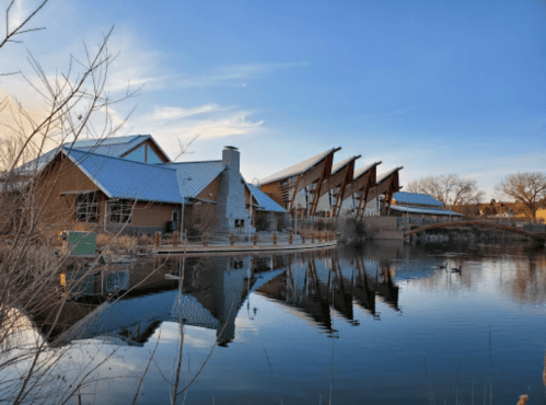 A modern building with a sloped roof reflects in a calm lake, surrounded by trees and a clear blue sky.