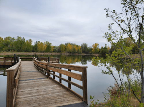A wooden dock extends into a calm lake, surrounded by trees with autumn foliage under a cloudy sky.