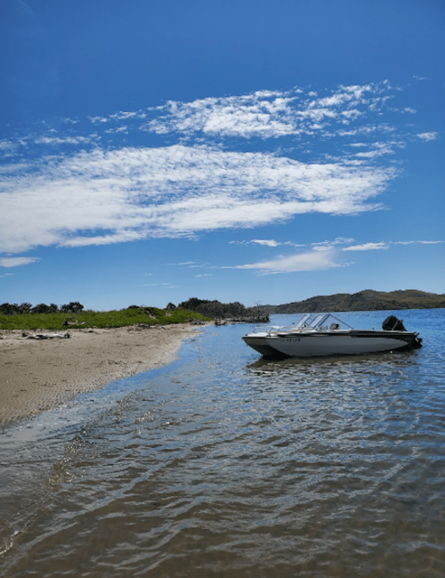 A small boat anchored near a sandy shore under a bright blue sky with scattered clouds.
