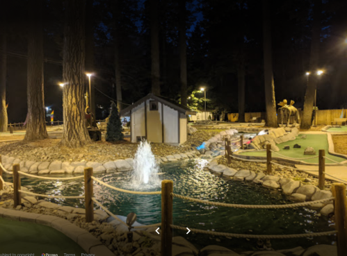 A nighttime view of a mini-golf course featuring a fountain, surrounded by trees and illuminated by soft lights.