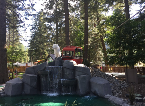 A bear statue stands on a rock formation near a small waterfall, surrounded by trees and a red gondola in the background.