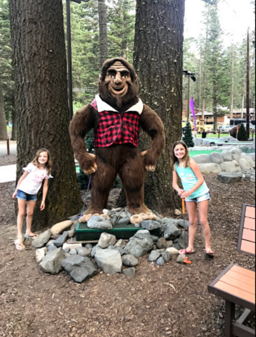 Two girls pose beside a large bear statue wearing a flannel shirt, surrounded by trees and rocks in a park setting.