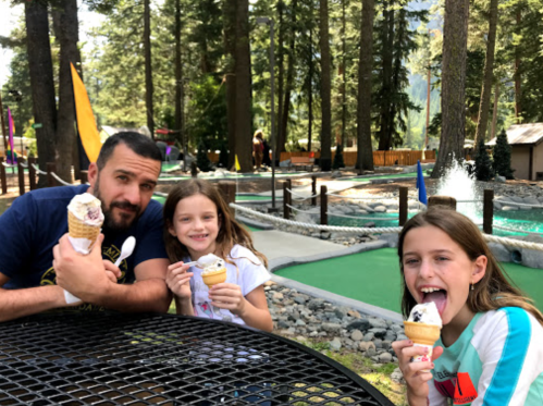 A man and two girls enjoy ice cream cones at a park with trees and a mini-golf course in the background.