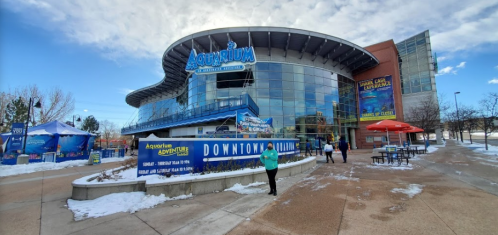 Exterior view of the Aquarium with a curved roof, surrounded by snow, featuring colorful signage and outdoor seating.
