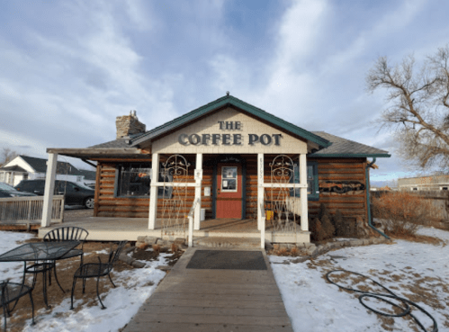 A rustic coffee shop with a wooden exterior, sign reading "The Coffee Pot," surrounded by snow and a clear blue sky.
