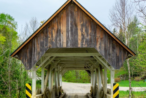 A wooden covered bridge with a peaked roof, surrounded by greenery and a gravel road leading through it.