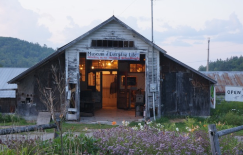A rustic barn with a sign reading "Museum of Everyday Life," surrounded by flowers and a cloudy sky.