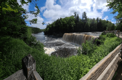 A scenic view of a waterfall surrounded by lush greenery and a cloudy blue sky, with a wooden railing in the foreground.