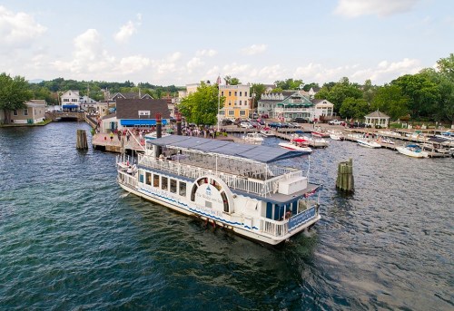 A riverboat docked near a waterfront town, surrounded by boats and buildings under a partly cloudy sky.