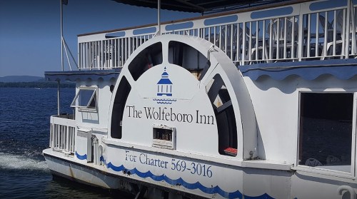 A boat named "The Wolfeboro Inn" with a paddle wheel, docked on a lake with mountains in the background.