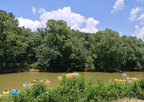 A river scene with colorful inflatable rafts floating among lush green trees under a bright blue sky with fluffy clouds.