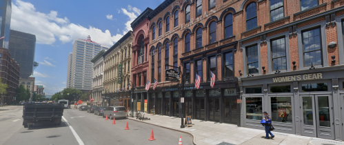 Historic brick buildings line a city street, with flags displayed and a woman walking past a women's clothing store.
