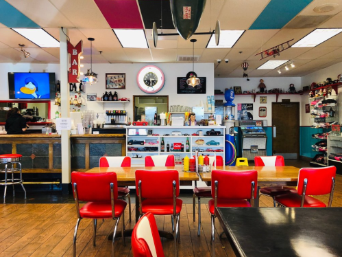 A retro diner interior with red chairs, a bar area, vintage decor, and a counter displaying drinks and snacks.
