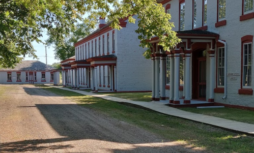 A gravel path leads past historic buildings with red trim and large windows, surrounded by trees and greenery.