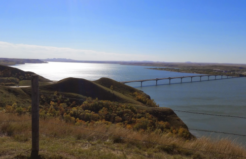 A scenic view of a river with a bridge, surrounded by rolling hills and autumn foliage under a clear blue sky.