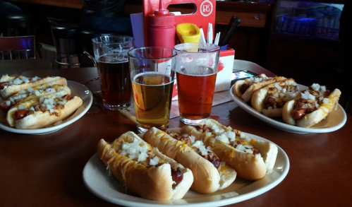 A table with four loaded hot dogs topped with onions and cheese, alongside two glasses of beer.