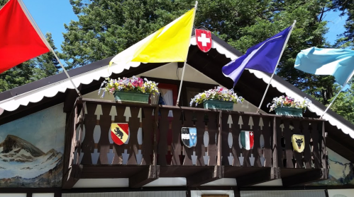 A wooden balcony adorned with colorful flags and flower pots, featuring painted shields and a mountain backdrop.
