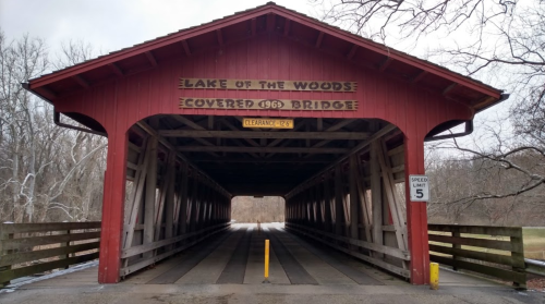Red covered bridge with "Lake of the Woods Covered Bridge" sign, surrounded by trees and a gravel path.