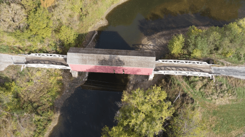 Aerial view of a red covered bridge spanning a calm river, surrounded by trees and autumn foliage.