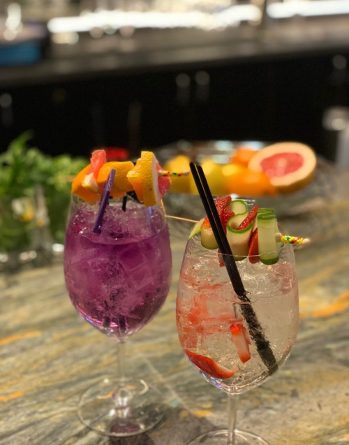 Two colorful cocktails on a marble countertop, one purple with fruit garnishes and one clear with cucumber and strawberries.