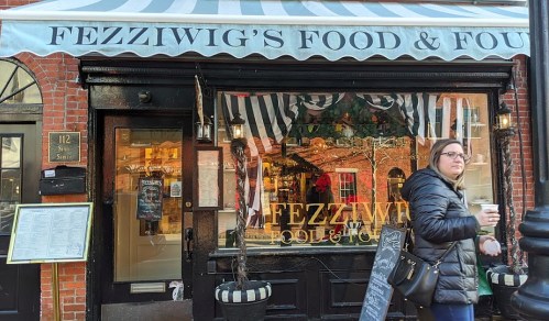 A woman walks by Fezziwig's Food & Fun, a cozy restaurant with a striped awning and festive decorations.