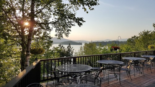 A scenic view from a deck with tables, overlooking a lake and mountains at sunset, surrounded by trees.
